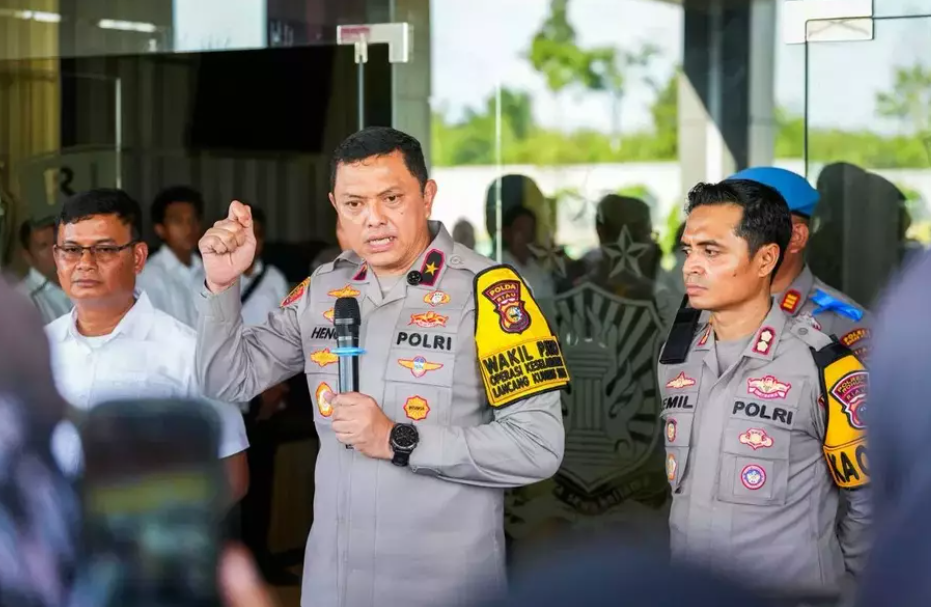 Perwira polisi menyampaikan keterangan pers di depan gedung sambil memegang mikrofon, didampingi anggota polisi lain dan sejumlah orang di belakangnya.