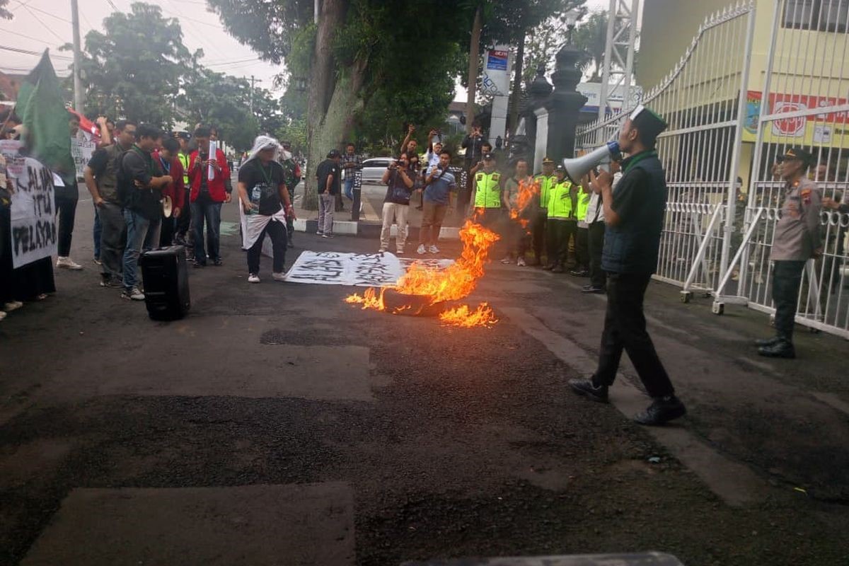 Sejumlah massa melakukan aksi unjuk rasa di depan gerbang gedung sambil membakar benda di tengah jalan, dengan pengamanan aparat kepolisian di sekitar lokasi.